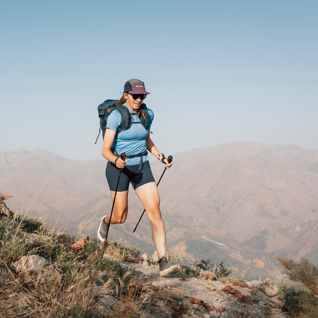 A woman hiking on a mountain ridge wearing a blue t-shirt, black shorts, a dark cap, and a black backpack with trekking poles, with a vast mountain range and clear blue sky in the background.