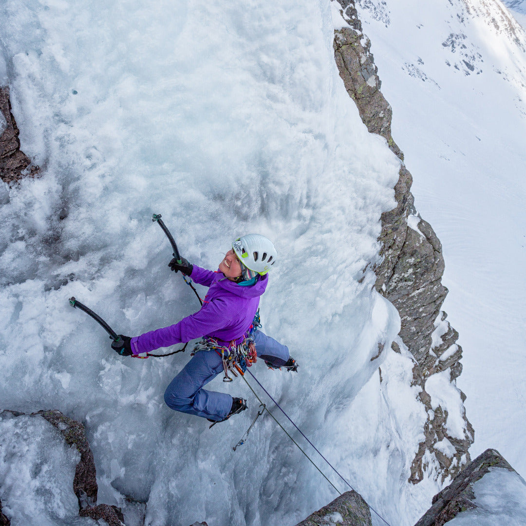 An ice climber wearing a purple jacket, blue pants, and a white helmet is suspended on a frozen cliffside, holding ice axes with ropes attached. The scene is set in a snowy mountain environment.