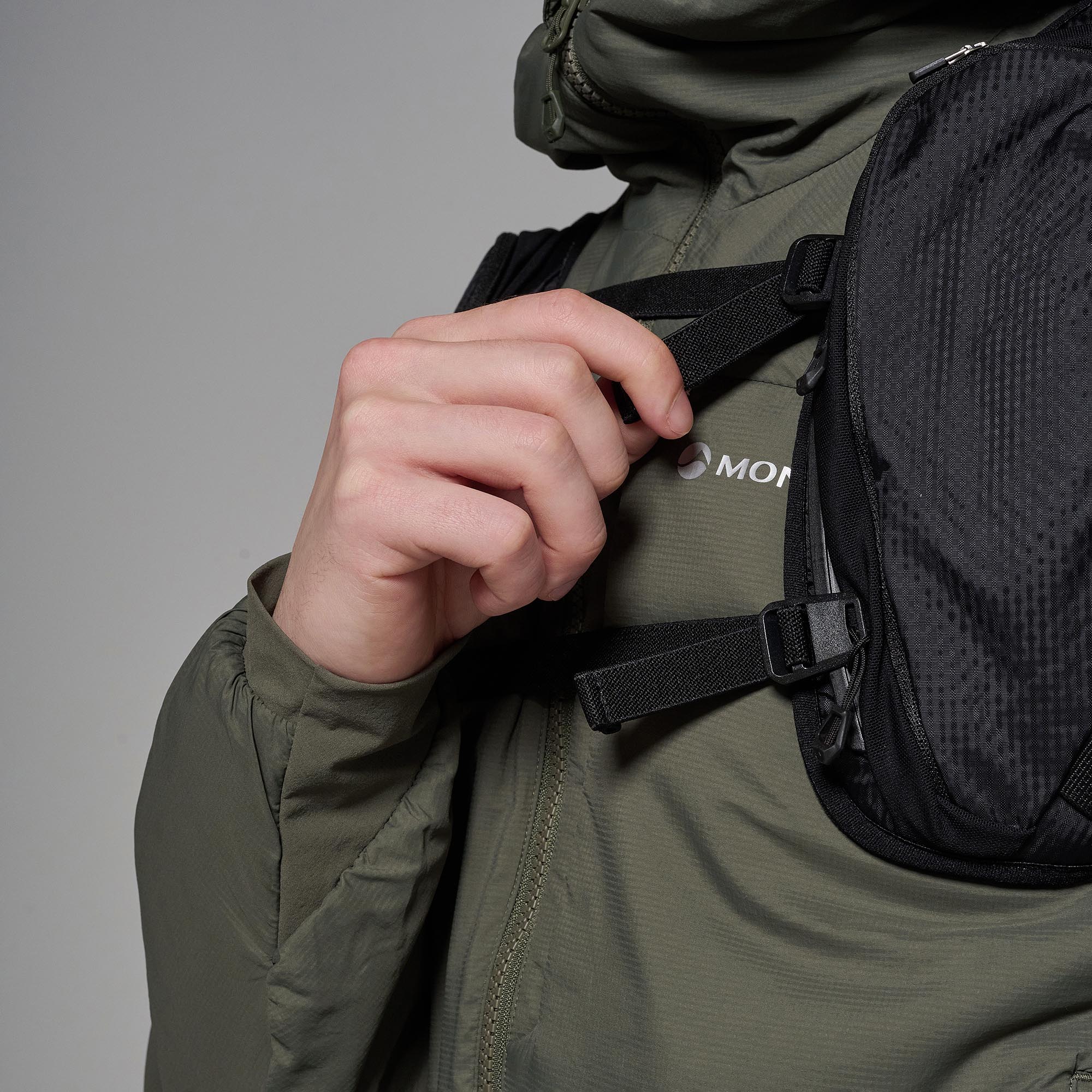 Close-up of a person wearing an olive green technical jacket and a black mesh backpack, adjusting a black strap on the chest. The brand logo 'MONT' is partially visible on the chest.