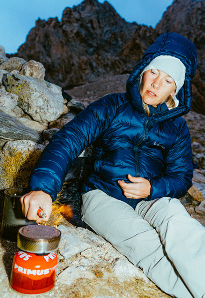 Woman lighting portable stove in down jacket sat against a rock
